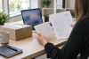 Une personne vérifie deux documents près d’un colis fermé et d’un écran flouté